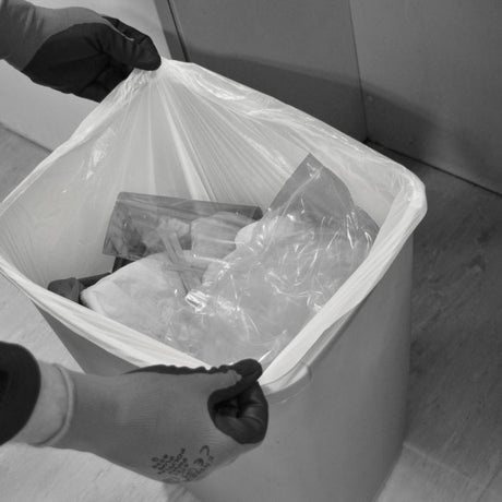Person wearing gloves holds a white trash bag over a bin filled with waste.